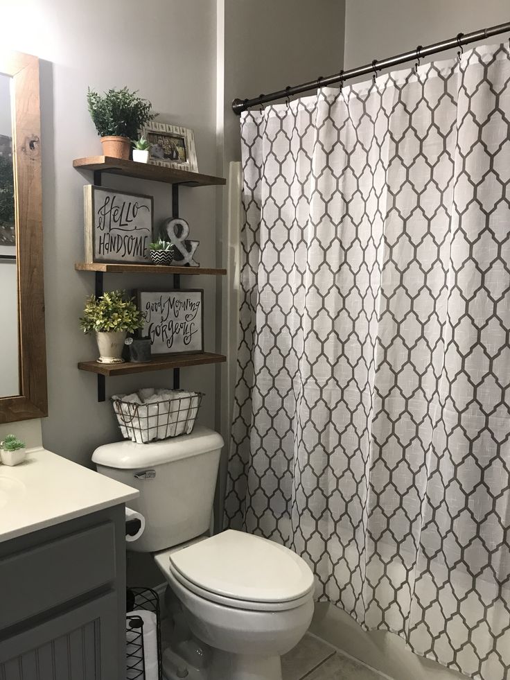 a white toilet sitting next to a shower curtain in a bathroom under a mirror on top of a wooden shelf