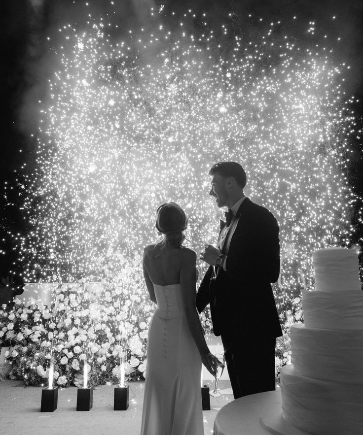 a man and woman standing next to each other in front of a cake with fireworks