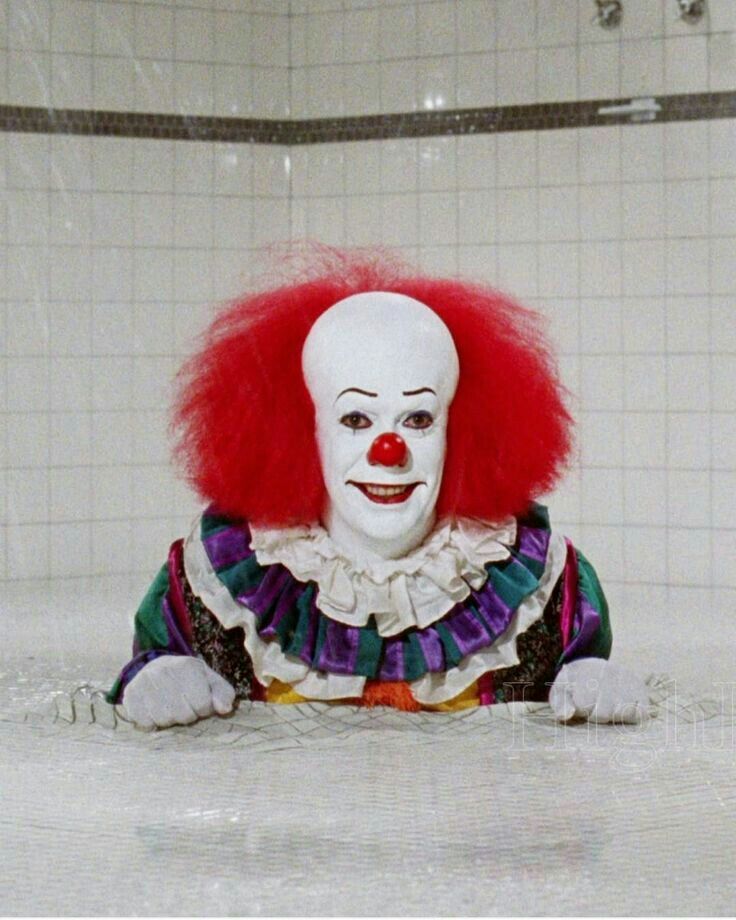 a clown sitting on the ground in front of a white tiled wall with red hair