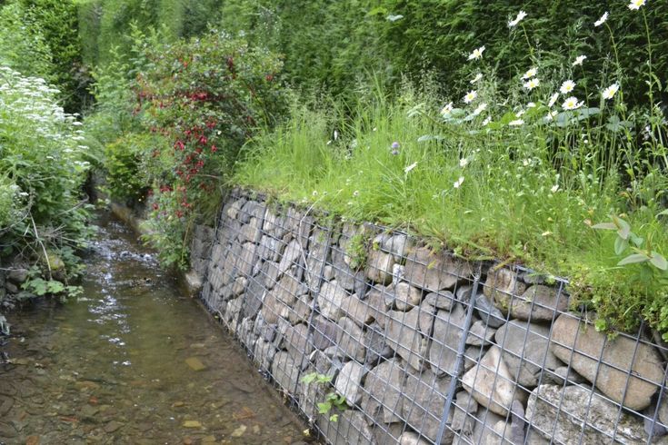 a stream running through a lush green forest next to a stone wall covered in flowers