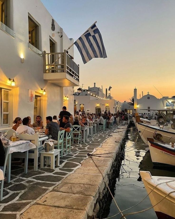 people sitting at tables in front of a building with boats parked on the water near by