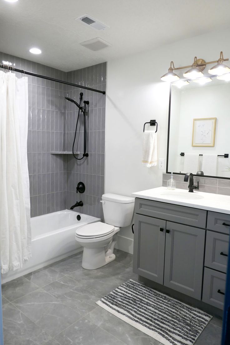 A bathroom with a tub/shower combo that has tub to ceiling gray glass subway tile installed vertically. The toilet is between the tub and single-sink vanity, which has darker gray cabinets and white Quartz countertops. The floor has large rectangular tiles that are gray with white veins. White Bathroom With Grey Floor, White And Dark Grey Bathroom, Bathroom Gray And Black, Grey Bathroom Styling, Modern Bathroom Grey And White, Grey Tile Shower Bathroom, Bathroom Remodel Gray And White, Small Bathroom With Grey Vanity, Small Bathroom Ideas Gray Vanity