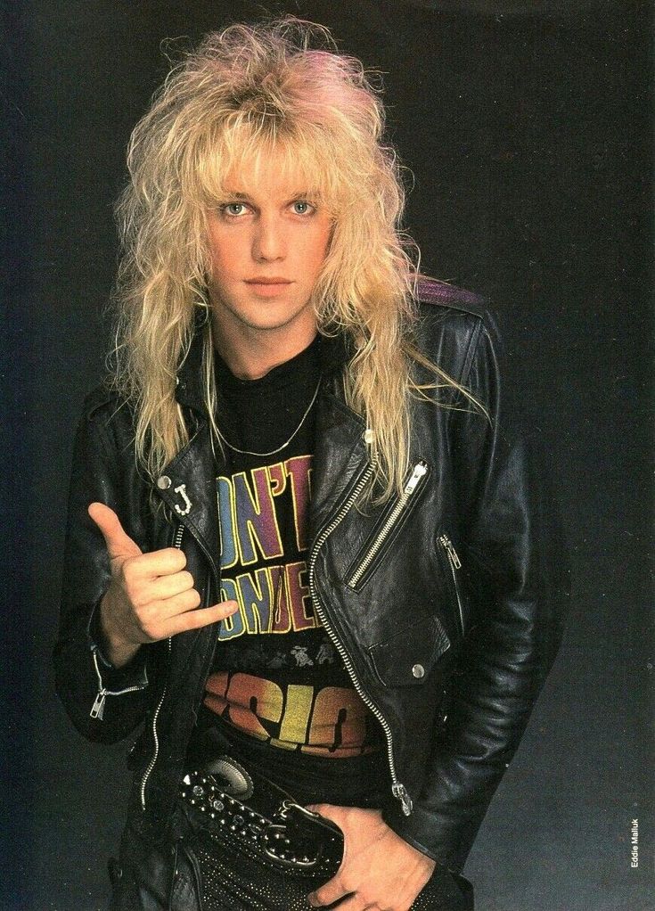 a man with long blonde hair wearing a black leather jacket and holding his finger up