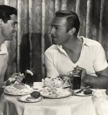 two men sitting at a table with food in front of them and one holding a drink