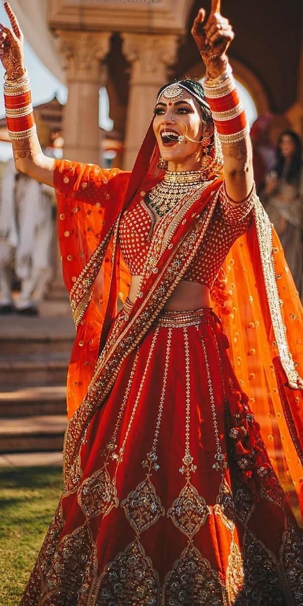 a woman in an orange and red bridal outfit dancing on the grass with her hands up