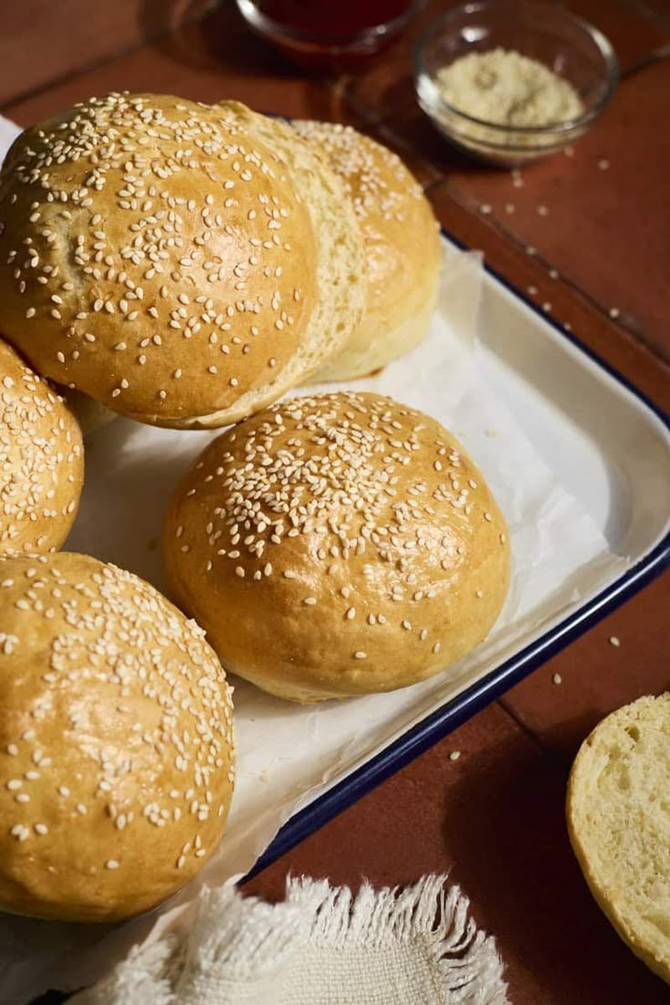 four sesame buns on a white plate next to some bread and other food items