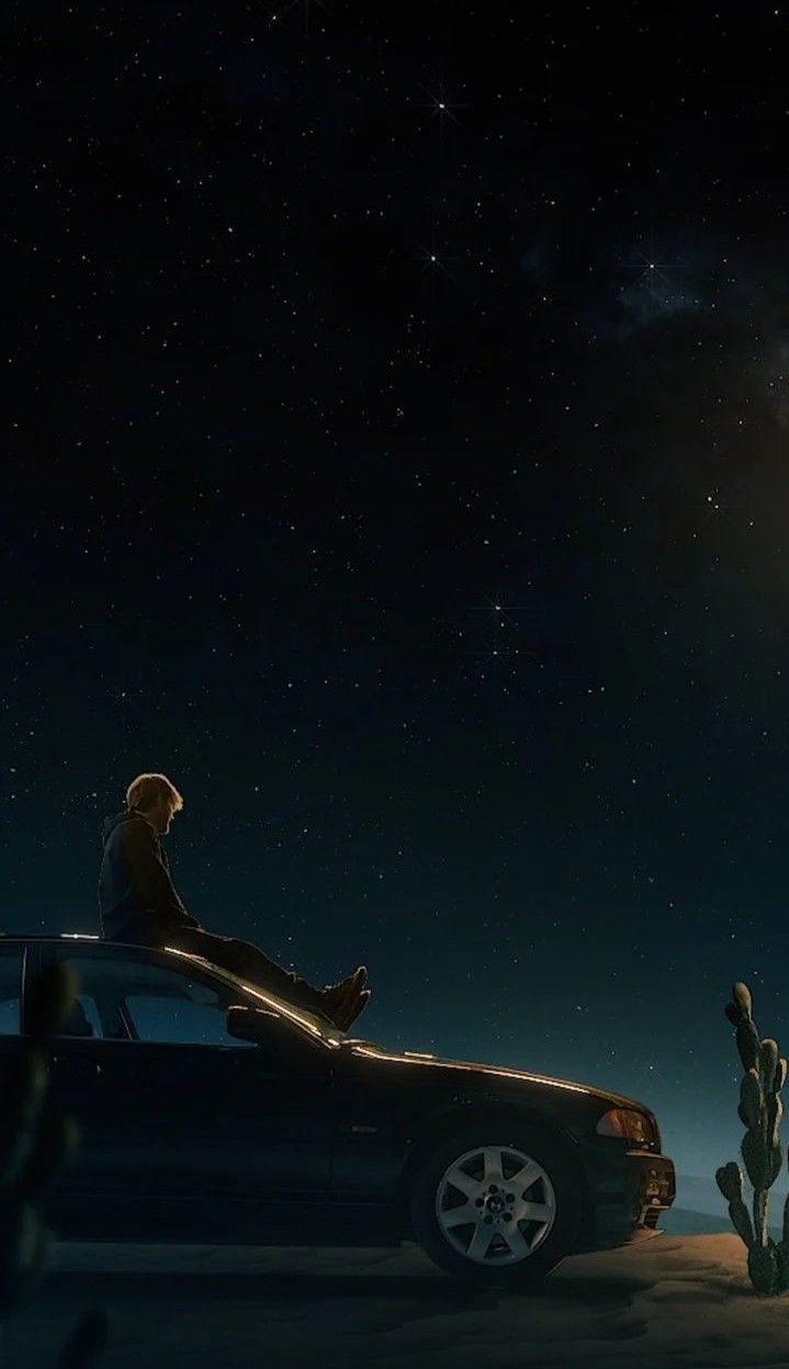 a man sitting on top of a car in the desert at night with stars above