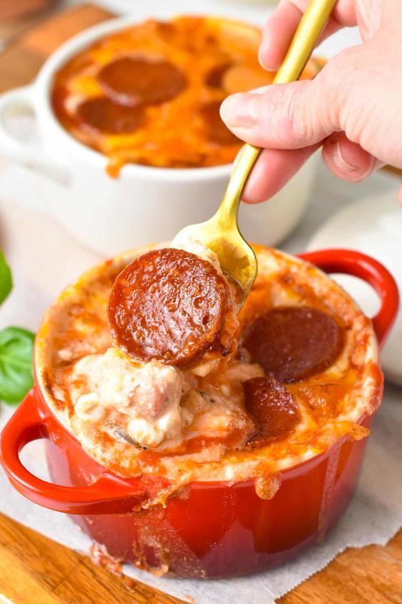 a spoon full of pizza dip being lifted from a red casserole dish with pepperoni and cheese