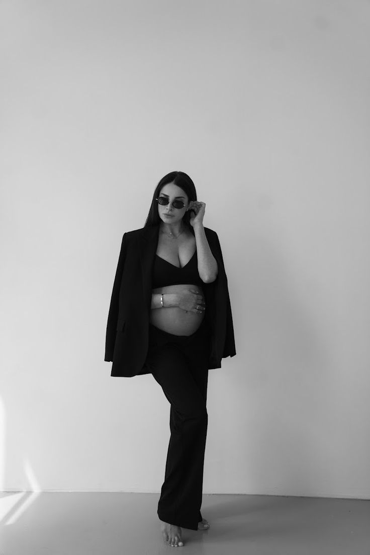 a black and white photo of a pregnant woman wearing sunglasses, standing in front of a wall