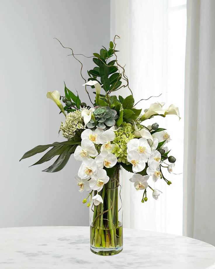 a vase filled with white flowers and greenery on top of a marble table next to a window