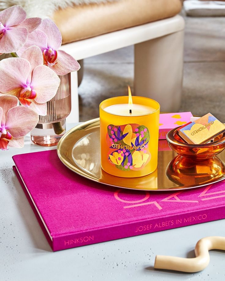 a yellow candle sitting on top of a table next to a pink book and some flowers