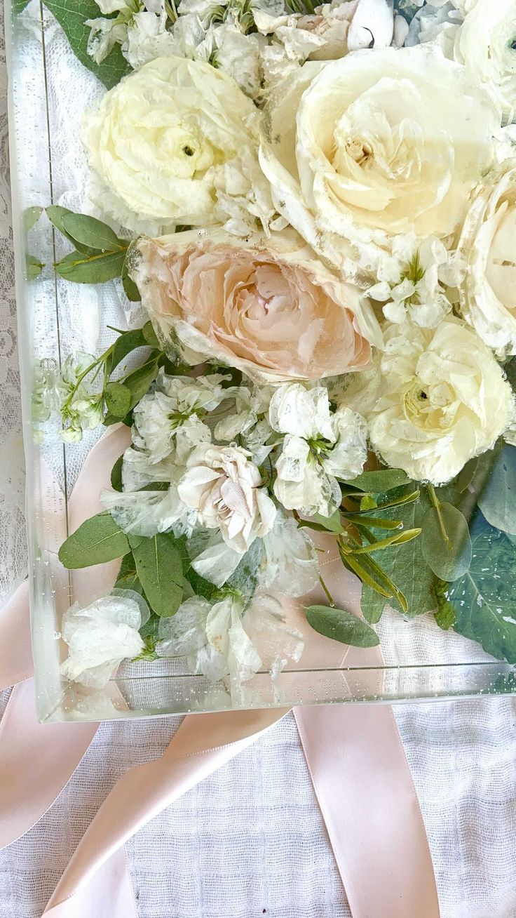 an arrangement of flowers is displayed in a clear box with pink ribbon on the side