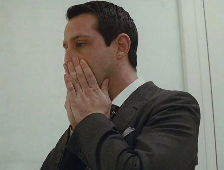 a man in a suit covers his face with his hands while looking into the mirror