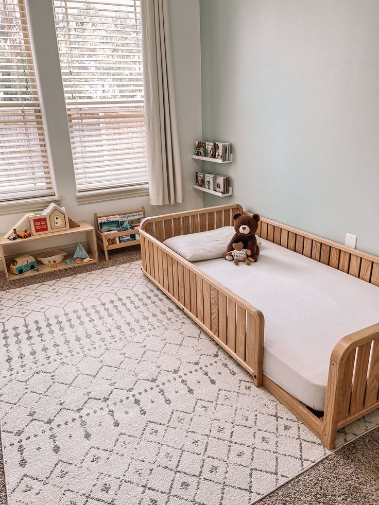 a baby crib with a teddy bear sitting on top of it in front of two windows