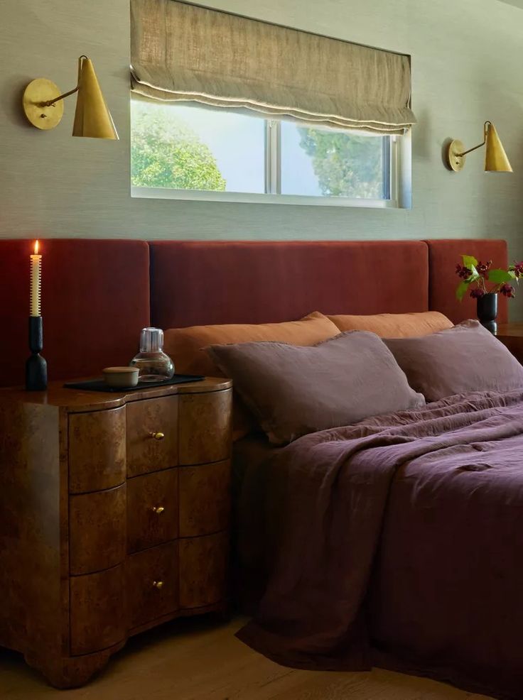 a bed with purple sheets and pillows in a bedroom next to a dresser, lamp and window