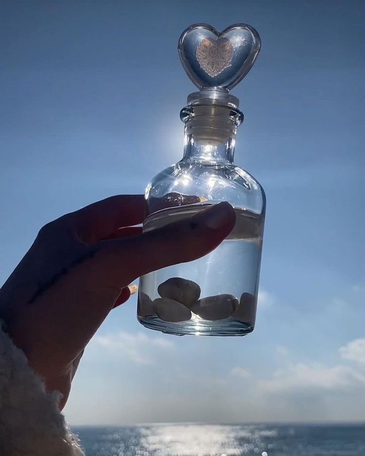 a person holding up a small bottle with rocks in it on the beach near the ocean