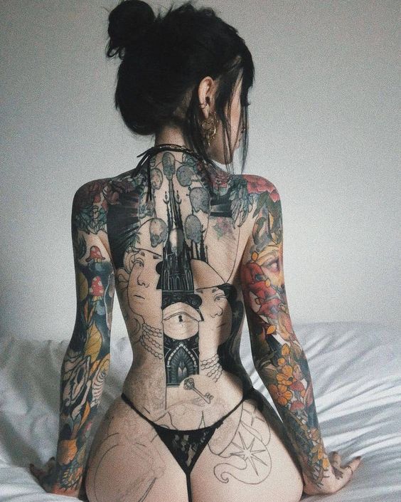 a woman with tattoos on her back sitting on top of a white bed covered in sheets