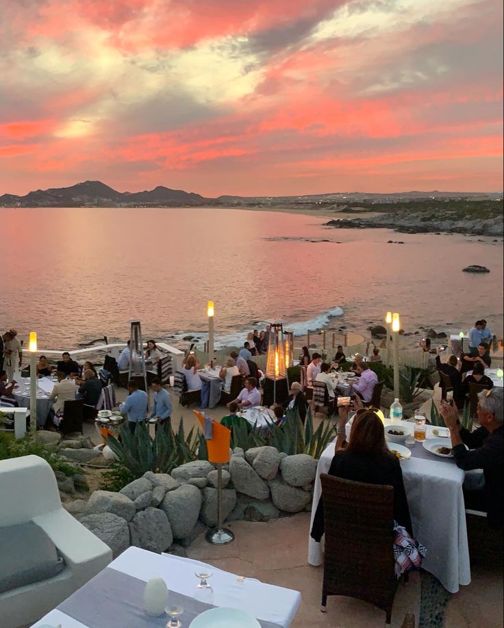 a group of people sitting at tables next to the ocean with candles in their hands