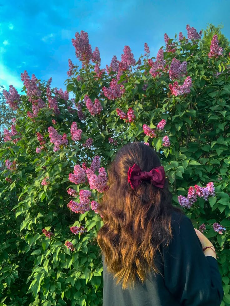 a woman with long hair and a bow tie standing in front of purple lilacs