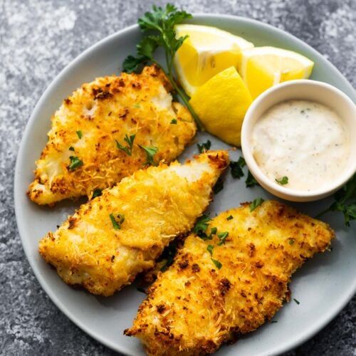 fried fish with lemon and parsley garnish on a white plate next to dipping sauce