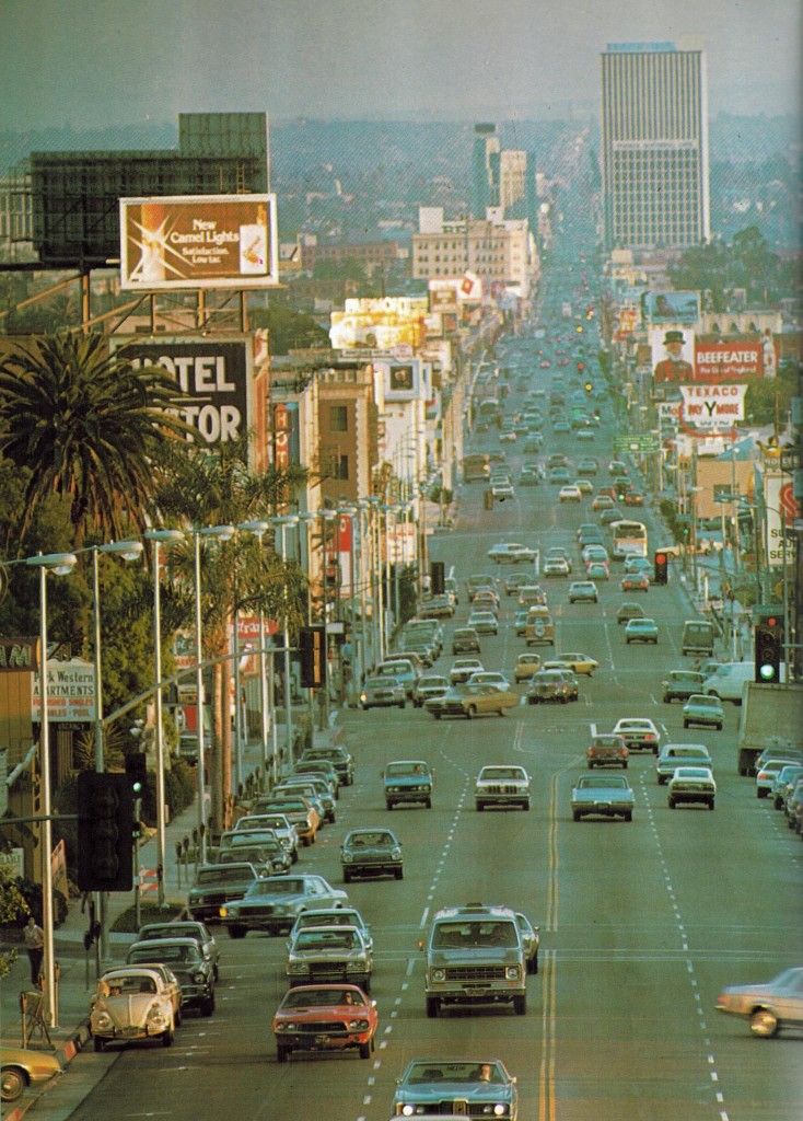 a city street filled with lots of traffic next to tall buildings and palm trees in the distance