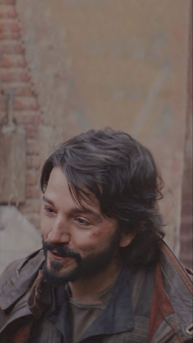 a man with long hair and beard standing in front of a brick wall wearing a leather jacket