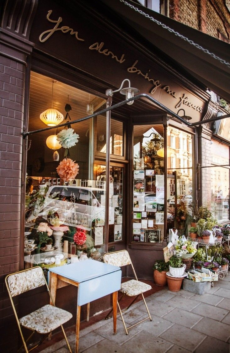 an outside view of a flower shop with many flowers in the window and chairs on the sidewalk