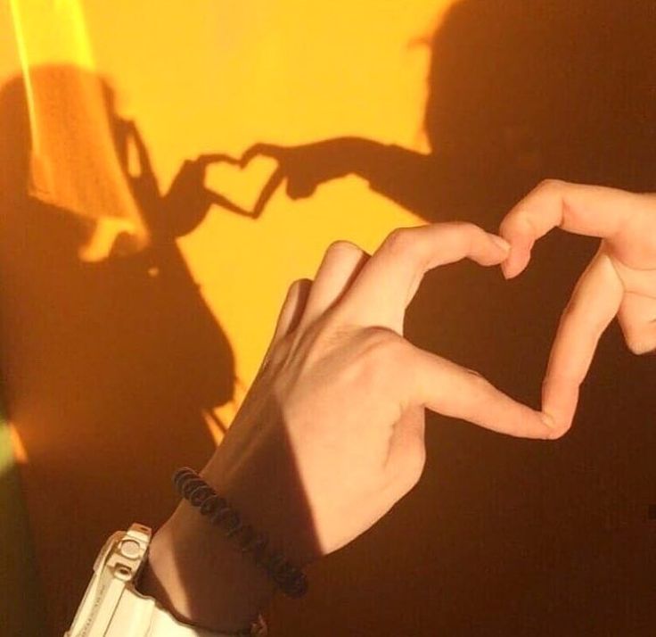 two hands making a heart shape with their fingers against a yellow wall and shadows cast on the wall