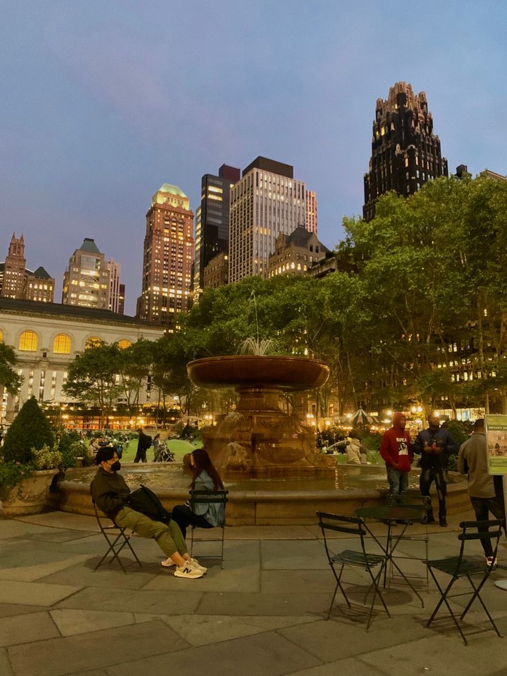 people are sitting in chairs near a fountain at night with skyscrapers in the background