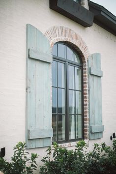 an open window with green shutters in front of a white brick wall and shrubbery