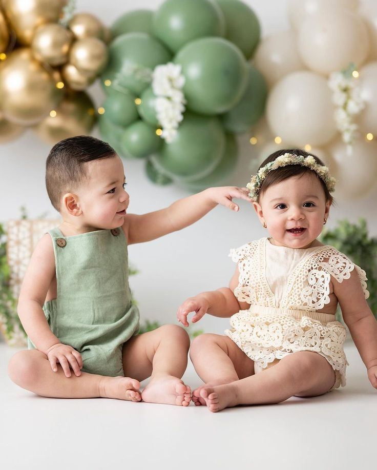 two babies sitting next to each other in front of balloons and streamers with gold, green and white decorations