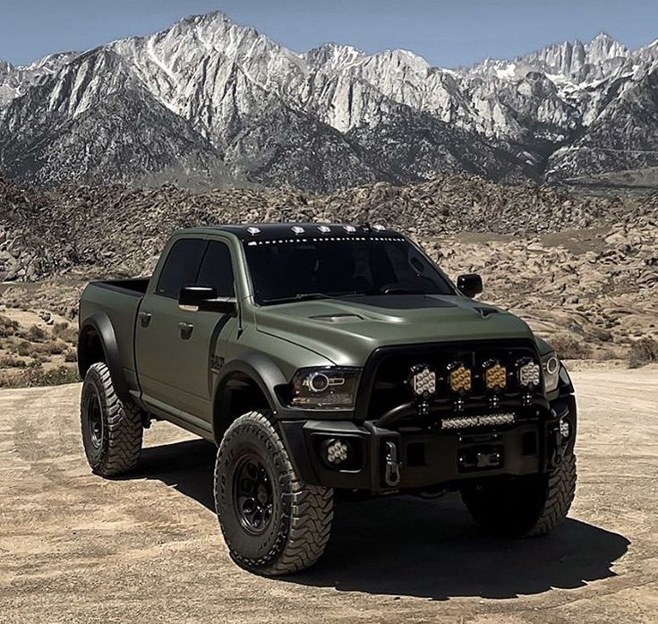 a large green truck parked on top of a dirt covered field in front of mountains