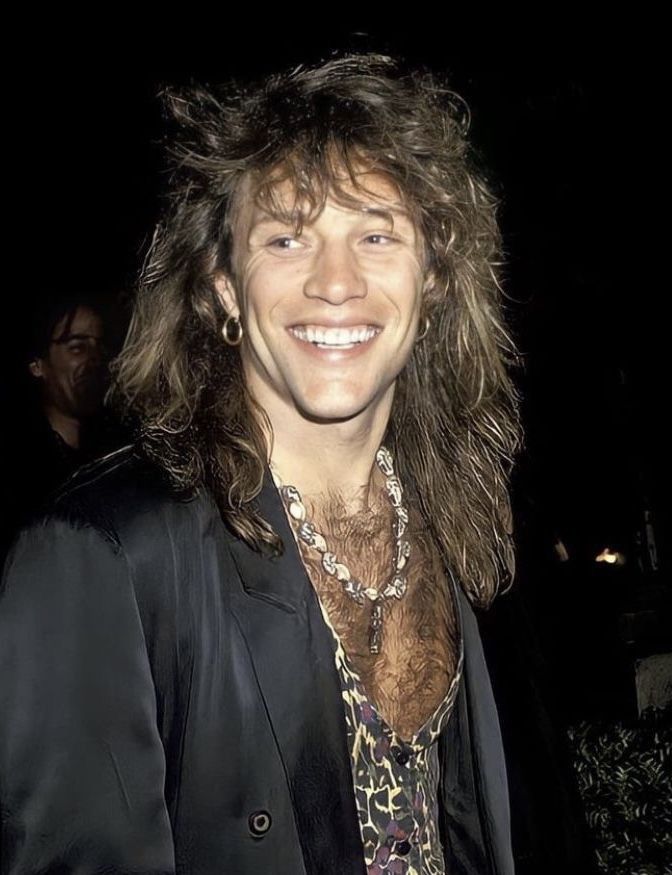 a man with long hair wearing a black jacket and leopard print shirt smiling at the camera