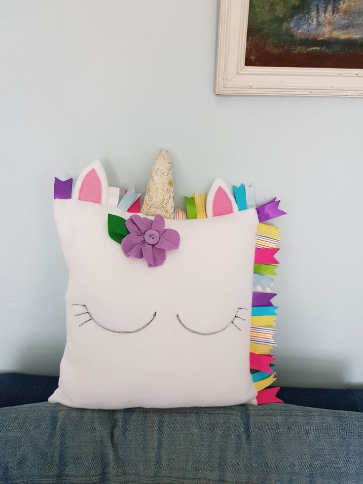 a white pillow with a unicorn face and colorful fringes on the front sitting on a blue couch