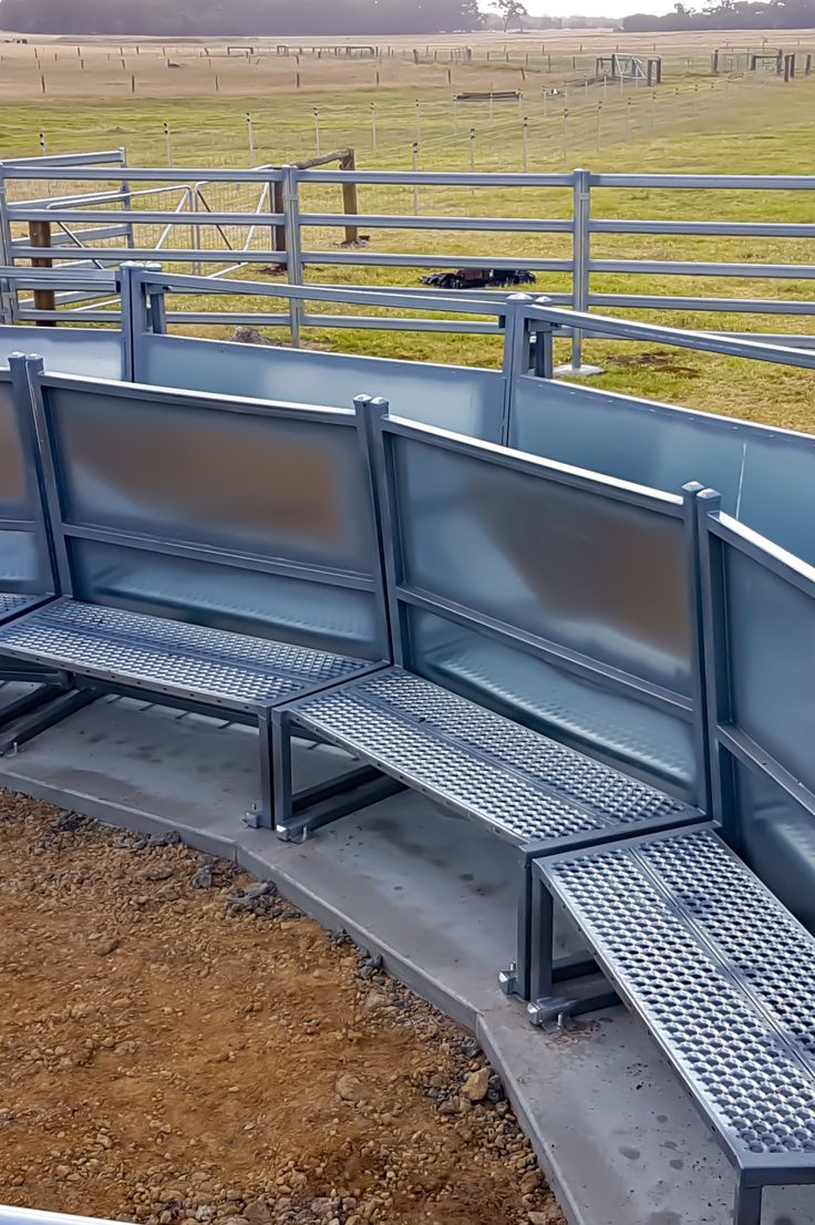 three metal benches sitting next to each other near a fenced in area with cattle