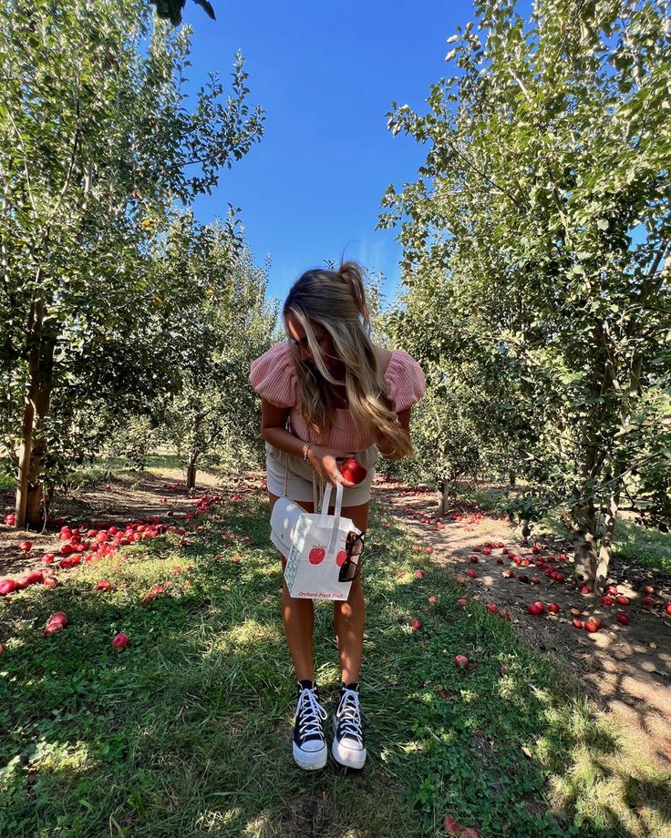 a woman standing in an apple orchard picking apples from the trees with her handbag