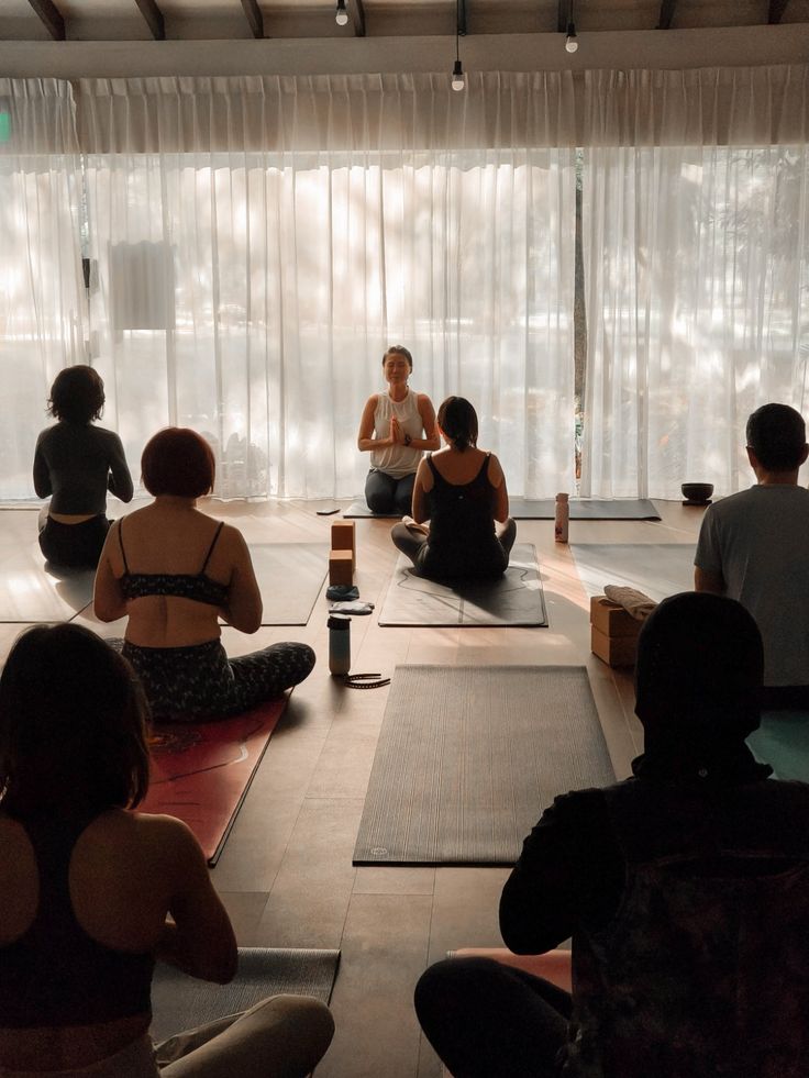 a group of people sitting on yoga mats in a room with large windows and curtains