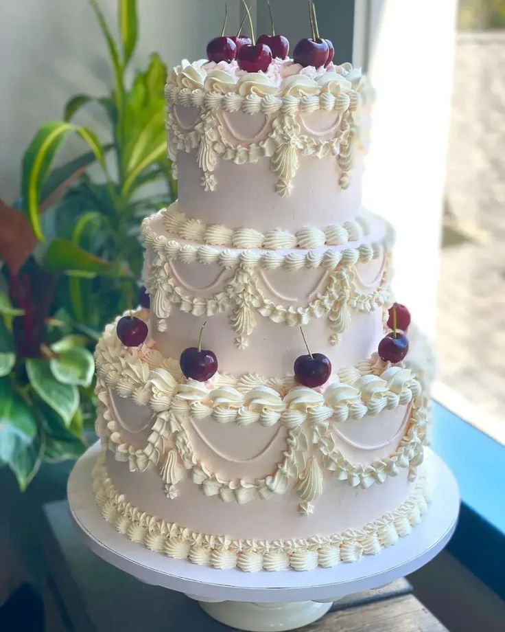 a three tiered wedding cake with cherries on the top and sides, sitting in front of a window