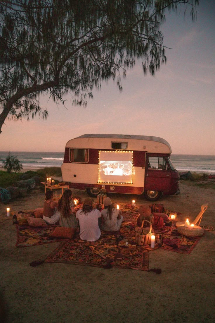 three people sitting on a rug in front of a camper van at the beach