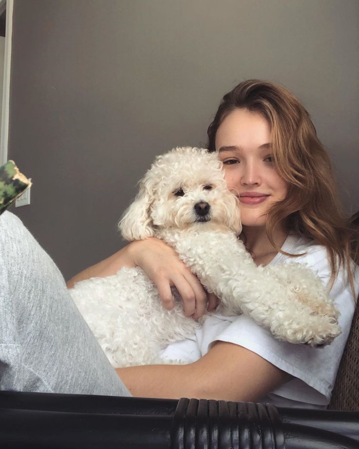 a woman holding a white dog in her arms while sitting on a couch next to a window