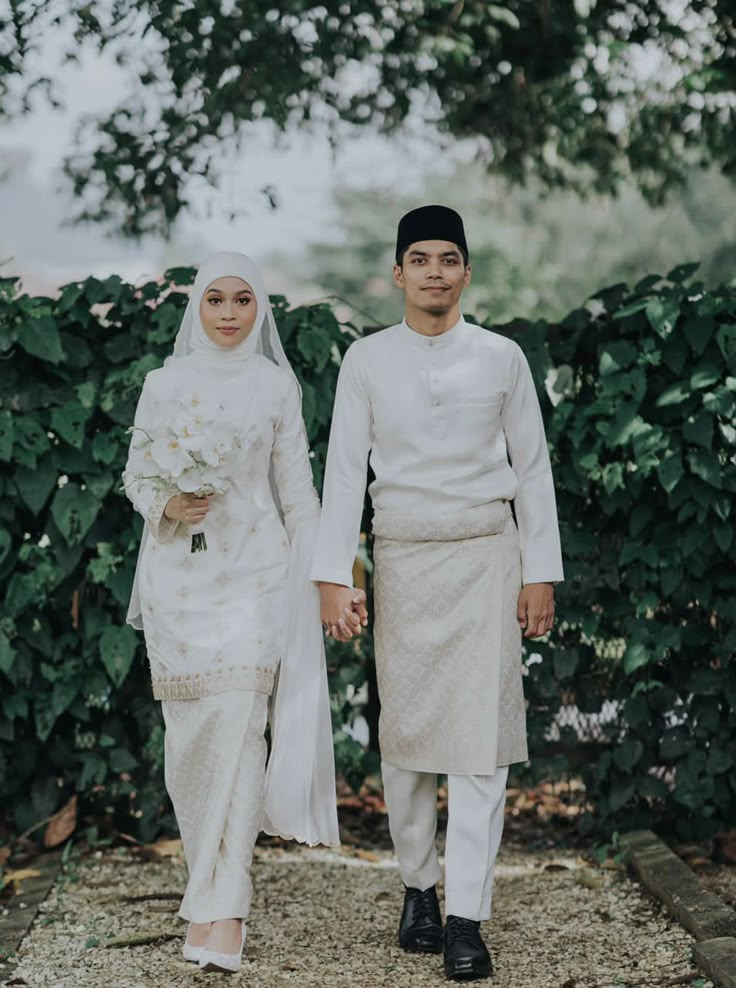 a man and woman dressed in white are holding hands while walking down a path with greenery behind them