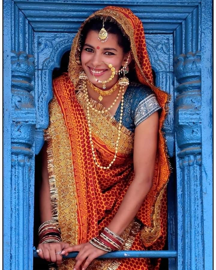 a woman in an orange and gold sari standing by a blue door with jewelry on her head