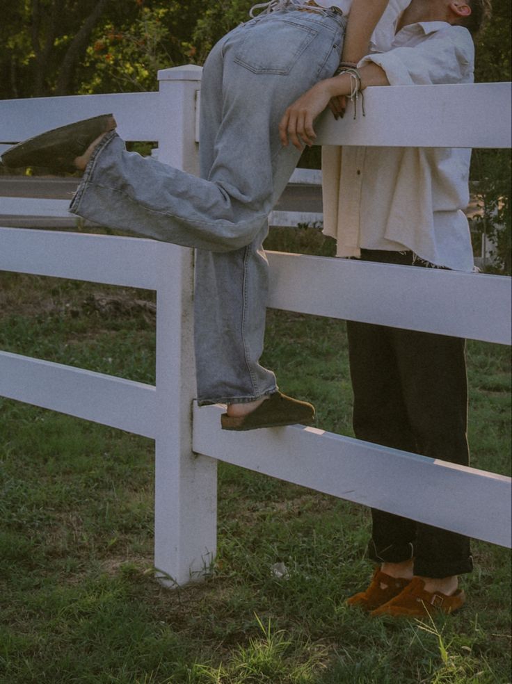 two people sitting on top of a white fence next to each other in the grass