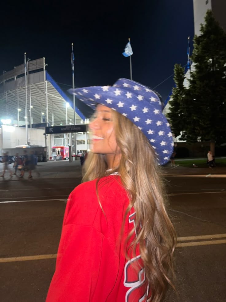 a woman with long hair wearing a red shirt and blue cowboy hat at night in front of an arena