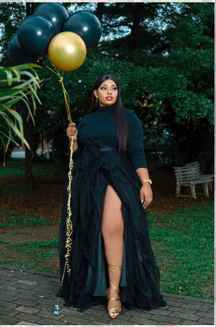 a woman in a black dress is holding some gold and green balloons while standing on a brick walkway