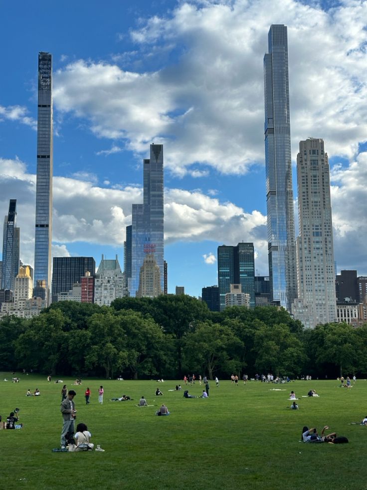 many people are sitting on the grass in front of some tall skyscrapers and trees