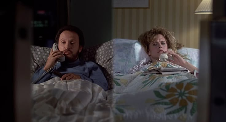 a man and woman laying in bed while talking on the phone, looking at each other