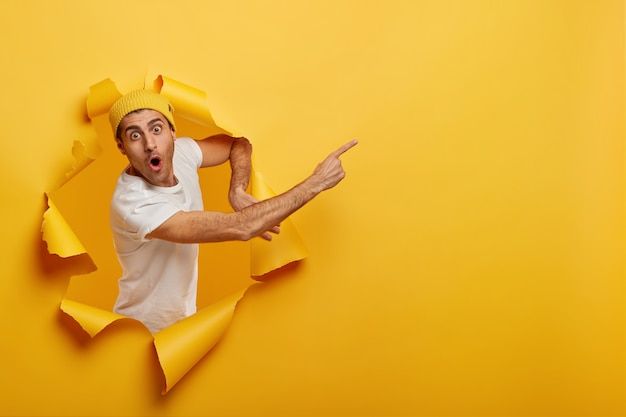 a man in white shirt and yellow hat making a face through torn up pieces of paper