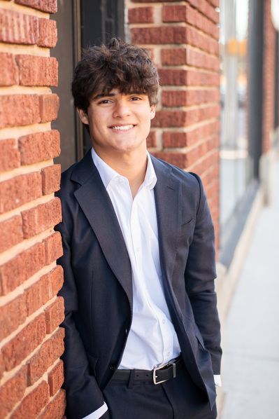 a young man leaning against a brick wall wearing a suit and white shirt with his hands in his pockets