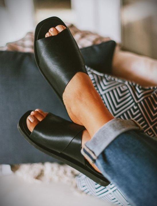 Close-up of a person's feet wearing Stormi Black Slide Sandals from Kay Kay Fashion. Their ankles are crossed and resting on a cushioned surface, showcasing women's flat shoes that are both comfortable and true to size. The person is wearing blue rolled-up jeans, with patterned cushions and soft furnishings in the background. Black Slides Women, Black Slide Sandals, Stylish Black Women, Black Work Shoes, Work Shoe, Chic Sandals, Black Slides, Latina Fashion, 2024 Style